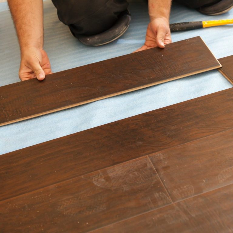 Person verlegt Holzfußbodenpaneele auf einem hellblauen Untergrund.